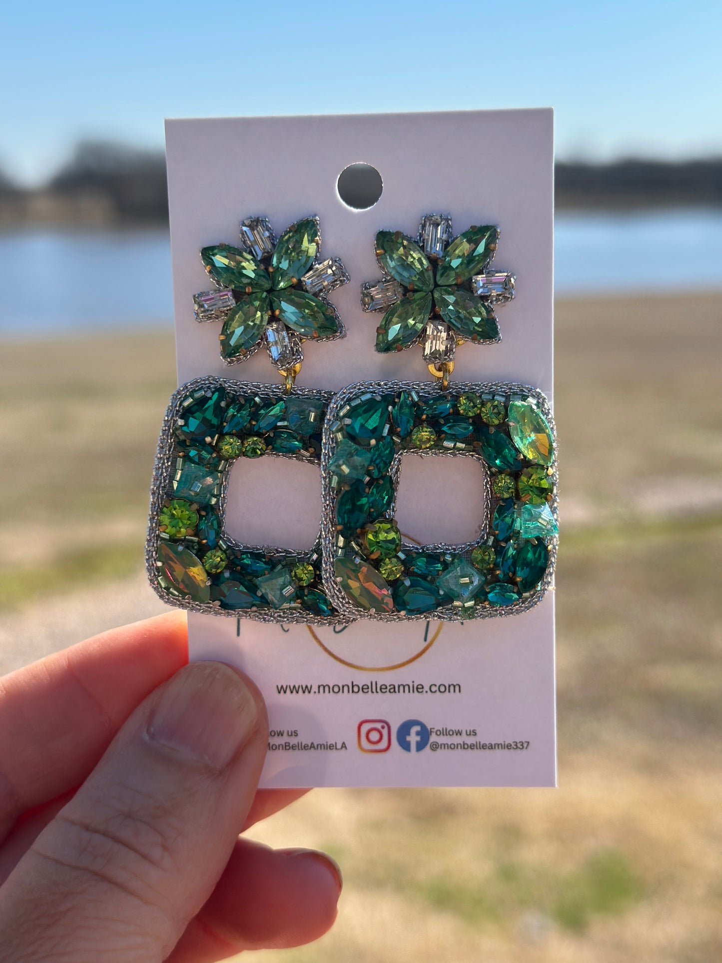 A pair of green and gold dangle earrings with gemstones and floral accents, displayed against an outdoor background.