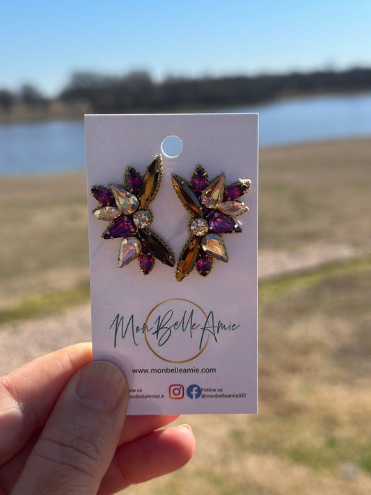 A pair of purple and gold gemstone stud earrings on a white card, held by a hand with a blurred background.