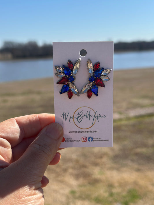 A pair of radiant cut gemstone stud earrings in red, white, and blue, held by a hand against an outdoor backdrop.